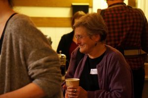 A photo of a woman holding a paper cup and smiling at somebody out of shot.