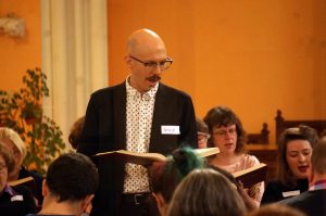 A man with a smart jacket and a moustache leads a song.