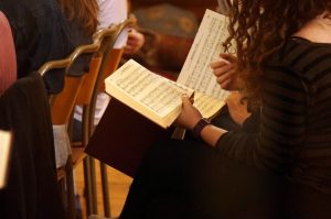 A photograph taken over the shoulder of a curly haired woman, showing her sacred harp book open on her lap. 