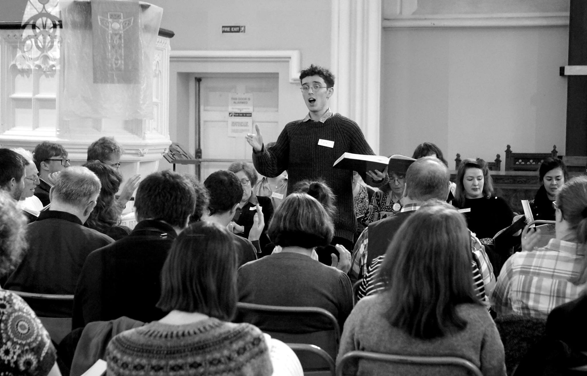 A black and white photograph of a young dark haired man, holding a songbook and raising his hand to lead a song. He is surrounded on all sides by seated singers facing towards him in a square formation. 