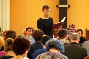 A man leads a song while holding a book, viewed from the tenor section. 