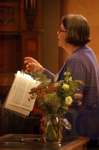 A side view of a woman singing and holding a book. She is standing beside a vase of flowers. 