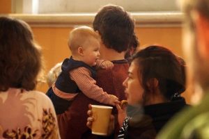 A photo of people mid conversation during the break between singing. A man is holding a baby on his shoulder and a woman is holding a paper cup.