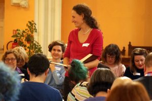 A woman with a red topand curly hair is smiling towards singers as she leads a song. 