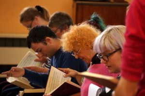 A side profile view of the front row of tenor singers. Several men and women sitting in a row singing from Sacred Harp books that are sitting on their knees. 