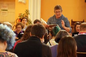 A woman in a striped top stands in the centre leading a song. 