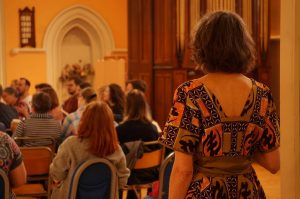 A photo from behind the alto section, a woman is standing away from the hollow square and wearing a colourful dress. She faces away from the camera towards the singers. 