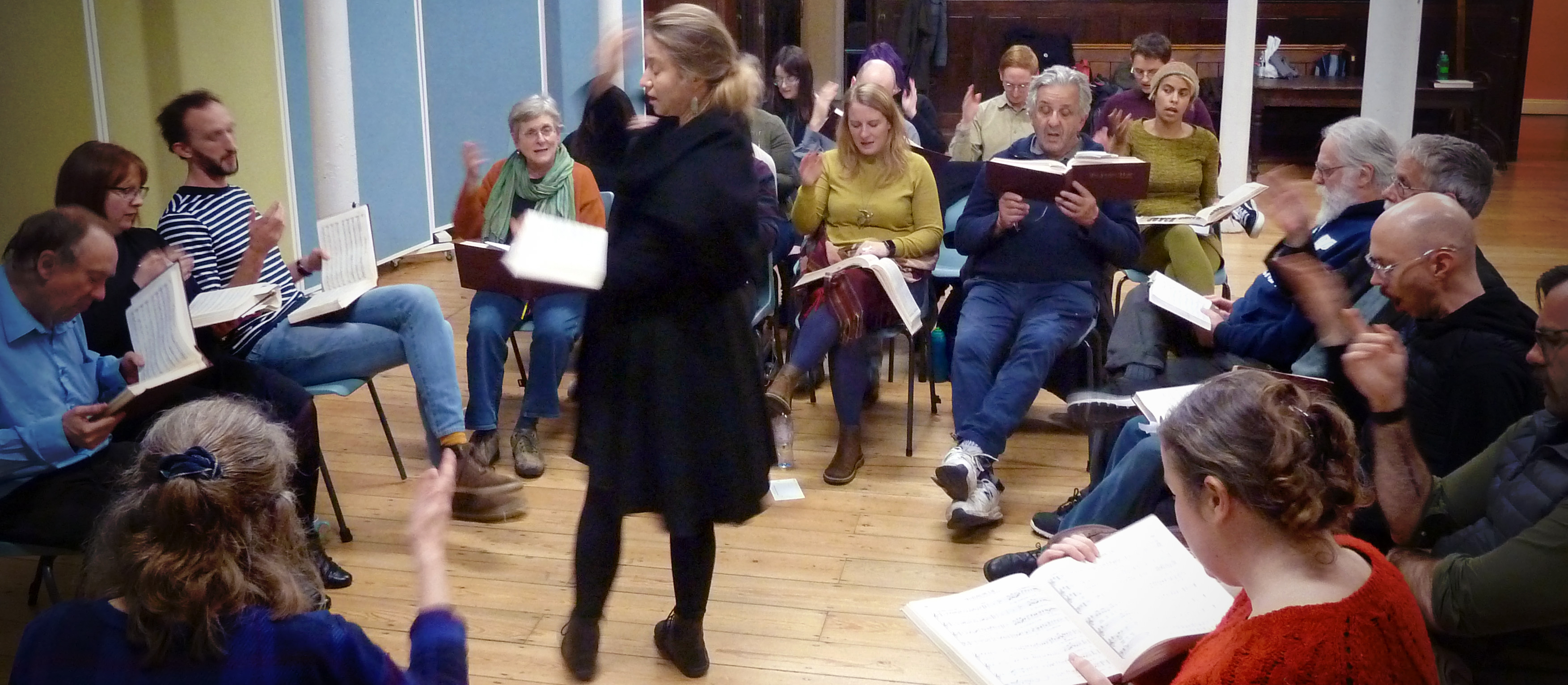 A woman standing, holding a songbook and raising her arm, leading a group of seated singers who are all facing towards her. 