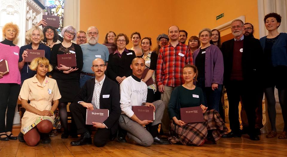 A group of about 20 people of varied age, ethnicity and gender are standing together in a church for a photograph. Many of them are holding a red songbook entitled "The Sacred Harp". Four of them are kneeling at in the front row.